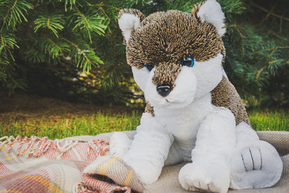 A soft, furry brown and white 16” plush wolf stuffy sitting on a blanket with green pines in background. *Comes unstuffed.